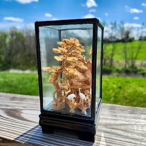 May include: A detailed wooden diorama of a pagoda, trees, and birds, encased in a glass box with a black frame. The miniature scene is set against a backdrop of green grass and a blue sky.