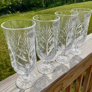 May include: Four clear glass goblets with a leaf pattern are arranged on a wooden deck. Each goblet has a wide, flared rim, a stem, and a round base. The goblets have a decorative leaf pattern etched into the glass. Each goblet has a small sticker that says "FIFTH AVE".