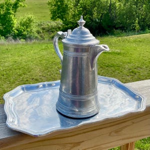 May include: A silver-colored metal coffee pot with a lid and a spout, sitting on a matching silver tray. The tray has a decorative, scalloped edge. The background shows green grass and trees.