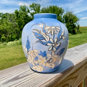 Könnte beinhalten: Eine hellblaue Keramikvase mit Craquelé-Glasur und handgemaltem Blumenmuster in Weiß und Gold. Das bündige Gefäß zeigt stilisierte gänseblümchenartige Blüten und Blätter, mit einem glatten, schmalen Hals und einem bauchigen, runden Körper.