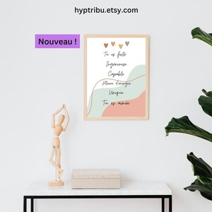 Może przedstawiać: Oprawiony wydruk artystyczny z jasną drewnianą ramą. Wydruk zawiera tekst "Tu es forte, Ingénieuse, Capable, Pleine d'énergie, Unique, Tu es aimée" w czarnym skrypcie, z pastelowymi akcentami kolorystycznymi. Słowo "Nouveau!" znajduje się w fioletowym pudełku.