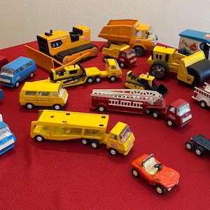 Puede incluir: Una colección de vehículos de juguete, incluyendo camiones, autobuses y equipos de construcción, en varios colores como amarillo, rojo, azul y blanco. Los juguetes están dispuestos sobre una superficie roja, mostrando una variedad de coches y camiones en miniatura.