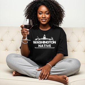 May include: A black t-shirt with the text "WASHINGTON NATIVE" in white, featuring a skyline graphic. The person is holding a glass of red wine and wearing grey sweatpants. The person is sitting on a cream-coloured sofa.