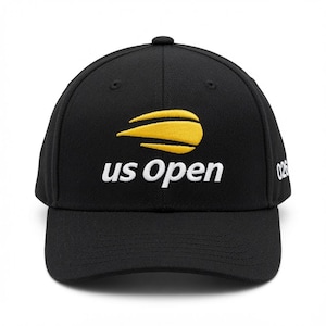 Op de afbeelding: Een zwarte honkbalpet met een geborduurde gele tennisbalafbeelding met een bewegingsspoor boven de witte tekst "us Open". Aan de zijkant van de pet staat het gedeeltelijke witte nummer "026". De pet heeft een gestructureerde kroon en een gebogen klep.