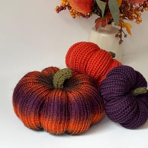 May include: Three crocheted pumpkins in different sizes and colours. One is a large orange and purple gradient, one is a medium solid orange, and one is a small solid dark purple. Each pumpkin features a small, textured green crocheted stem.