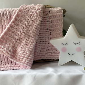 May include: A soft, light pink chunky knit blanket with a textured, looped yarn pattern. Beside the folded blanket sits a white star-shaped lamp featuring a printed face with closed eyes, a small smile, and pink circular cheeks, next to white flowers.