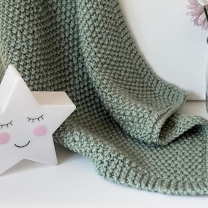 May include: A sage green knitted blanket is draped over a surface, next to a white star-shaped night light with a smiling face and closed eyes. A clear glass vase holds pink and white flowers.