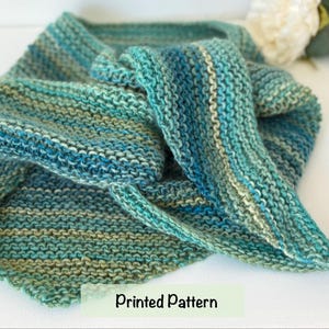 May include: A hand-knitted triangular shawl featuring a garter stitch texture with variegated yarn in shades of teal, turquoise, forest green, and olive. The fabric displays horizontal stripes of colour throughout the piece. Text reads: Printed Pattern.