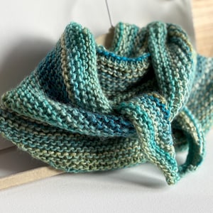 Op de afbeelding: Een handgebreide col sjaal in tinten van blauwgroen, blauw en groen. De sjaal is gemaakt van zacht, getextureerd garen en wordt getoond op een beige kleerhanger. Het gebreide ontwerp is zichtbaar, wat het vakmanschap van het stuk laat zien.