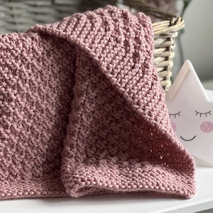 Può includere: Una coperta lavorata a maglia color rosa polvere con un motivo a legaccio testurizzato. Il tessuto è ripiegato su se stesso, mostrando punti a maglia uniformi su tutto il materiale. La coperta è posizionata accanto a una luce notturna a stella.