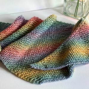May include: A collection of hand-knitted, colorful scarves. The scarves display a gradient of colors, including green, yellow, orange, pink, and purple. The knitted texture is visible, and the scarves are folded and arranged on a white surface.