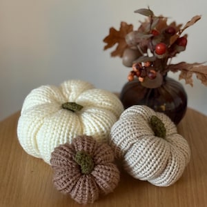 May include: Three crocheted pumpkins in cream, beige, and brown, each featuring a small green stem. The pumpkins are arranged together, with the largest cream one in the back, a medium beige one to the right, and a small brown one in the front.