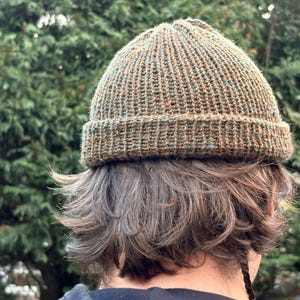 May include: A knitted beanie hat in shades of brown, green, and orange. The hat has a ribbed brim and a textured, close-knit crown. The hat is worn by a person with brown hair, with a green leafy background.