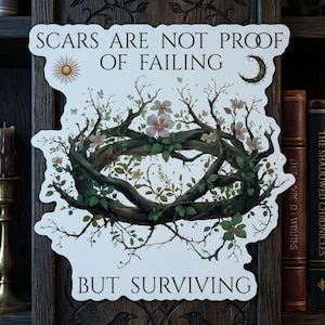 May include: A white sticker with the text "SCARS ARE NOT PROOF OF FAILING BUT SURVIVING" and a floral wreath design. The sticker has a sun and moon graphic. The background includes books, a candle, and a skull.