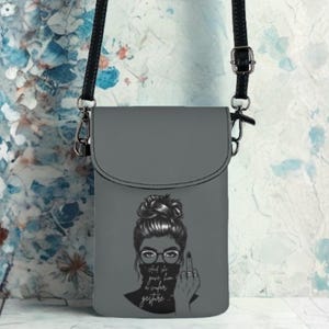May include: A gray crossbody phone bag with a black strap. The bag features a graphic of a woman with glasses and a bun, giving the middle finger. The text on the bag reads "and she goes, fuck you, maybe".