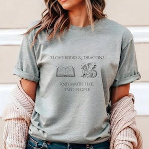 以下が含まれることがあります： 「I LOVE BOOKS & DRAGONS」の文字と、開いた本とドラゴンのイラストが描かれたライトグレーのTシャツ。イラストの下には「AND MAYBE LIKE, TWO PEOPLE」と書かれています。