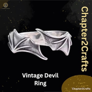 Może przedstawiać: Srebrny pierścionek z motywem skrzydeł diabła. Pierścionek posiada dwa szczegółowe kształty skrzydeł na górze, z napisem "Vintage Devil Ring" na dole. Tło jest czarne ze złotymi akcentami.