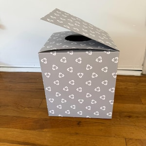 May include: A square cardboard box with a light gray finish and a repeating white pattern of recycling symbols. The box features a top flap that is partially open, revealing a circular opening on the upper surface of the container.