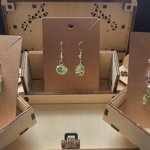May include: Three pairs of handmade earrings displayed on brown cardboard earring cards inside a wooden box. The earrings feature green glass beads wrapped in silver wire. The box is open, showcasing the earrings.