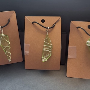 May include: Three necklaces with olive-green glass pendants wrapped in silver wire, each suspended from a black cord. The pendants are displayed on brown cardboard tags, creating a minimalist aesthetic. The pendants vary slightly in shape and wire wrapping style.