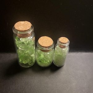 Può includere: Tre barattoli di vetro trasparente con tappi di sughero, ciascuno riempito con pezzi di vetro verde di forma irregolare. I barattoli variano in dimensioni, il più grande a sinistra e il più piccolo a destra. Lo sfondo è nero.