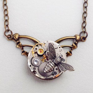 May include: A bronze chain necklace with a circular pendant featuring a detailed clockwork design and a silver bee. The pendant is attached to two bronze wings.