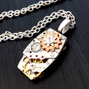 Peut inclure: Collier argenté avec un pendentif rectangulaire composé d'engrenages de montre et d'une chaîne argentée. Le pendentif comporte un engrenage cuivré avec un bijou central et de petits bijoux rouges. Le collier est sur une surface sombre.