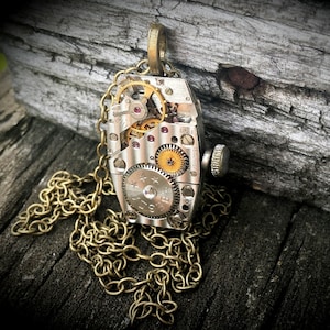 Peut inclure: Un collier en argent et or avec un pendentif présentant un design d'horlogerie détaillé. Le pendentif est de forme rectangulaire aux bords arrondis et présente des engrenages visibles et d'autres pièces d'horlogerie. Le collier est fait d'une chaîne en laiton.