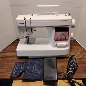 May include: A white Brother sewing machine with a top carrying handle, a red control panel displaying various stitch patterns, and a reverse button. Included are two blue fabric swatches with white stitching and a black foot pedal with a power cord.