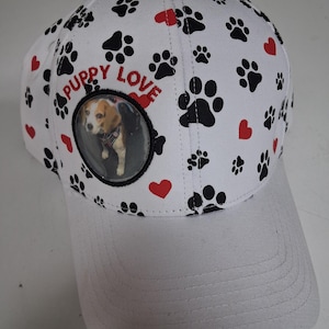 May include: White baseball cap with a curved brim, featuring a pattern of black paw prints and red hearts. The cap includes the text "PUPPY LOVE" and a circular image of a dog.