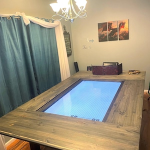 May include: A large, rectangular wooden table with a built-in illuminated grid. The table has a dark gray stain and sturdy legs. The center features a blue-lit grid, perfect for tabletop gaming. A chandelier hangs above the table.