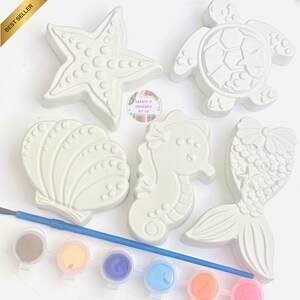 May include: A set of five white plaster sea-themed figurines including a starfish, turtle, seashell, seahorse, and mermaid tail. A blue paintbrush and a strip of six small paint pots containing various colors are included for painting the figures.