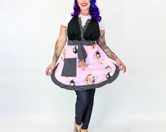 Op de afbeelding: Een roze en zwarte schort met een zwart stippenpatroon en een geruite zoom. Het schort heeft een grote zak aan de voorkant en heeft een print van vrouwen in vintage kleding.