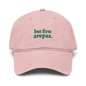 Könnte beinhalten: Hellrosa Baseballkappe mit gebogenem Schirm. Auf der Kappe steht der grüne Text "but first arepas" mittig auf der Vorderseite. Die Kappe besteht aus einem weichen Stoff und hat ein klassisches, verstellbares Design.