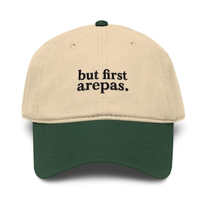 Könnte beinhalten: Baseballkappe mit cremefarbener Krone und dunkelgrünem Schirm. Der Text "but first arepas" ist in Schwarz auf die Vorderseite der Kappe gestickt. Die Kappe hat einen dunkelgrünen Knopf oben.