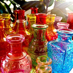 May include: A collection of colorful glass bottles in shades of red, yellow, green, and blue. The bottles have various shapes and textures, with some featuring embossed patterns. The arrangement creates a vibrant display, perfect for home decor.