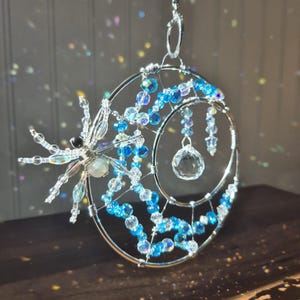 May include: A silver wire suncatcher with a crescent moon design. The moon is adorned with blue and clear beads, and a crystal ball hangs from the center. A wire spider with crystal accents is on the left side.