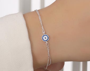 Puede incluir: Una pulsera de plata con un pequeño dije redondo con un diseño de ojo turco azul y blanco. La pulsera es delicada y tiene una cadena simple. La imagen sugiere que es una pieza de joyería.