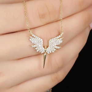 Puede incluir: Collar dorado con un colgante en forma de alas de &aacute;ngel. Las alas est&aacute;n adornadas con peque&ntilde;os cristales brillantes. El colgante cuelga de una fina cadena. El collar se muestra sobre un fondo oscuro.