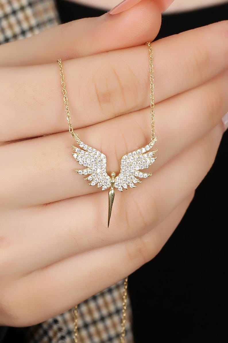 Puede incluir: Collar dorado con un colgante en forma de alas de &aacute;ngel. Las alas est&aacute;n adornadas con peque&ntilde;os cristales brillantes. El colgante cuelga de una fina cadena. El collar se muestra sobre un fondo oscuro.
