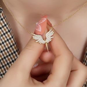 Puede incluir: Un collar dorado con un colgante en forma de &aacute;ngel con alas extendidas. Las alas y el cuerpo est&aacute;n incrustados con peque&ntilde;os cristales brillantes. El collar se sostiene, mostrando el dise&ntilde;o.