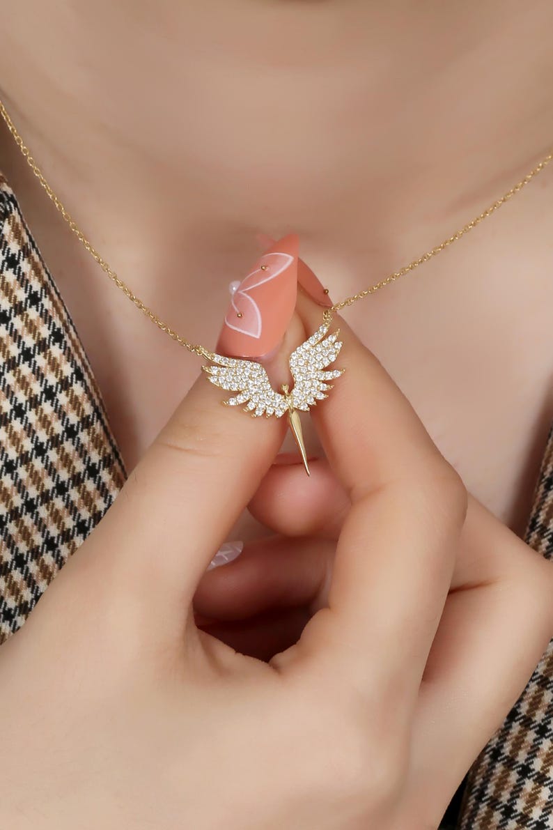 Puede incluir: Un collar dorado con un colgante en forma de &aacute;ngel con alas extendidas. Las alas y el cuerpo est&aacute;n incrustados con peque&ntilde;os cristales brillantes. El collar se sostiene, mostrando el dise&ntilde;o.