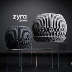 May include: Two gray, spherical planters with a geometric design, each sitting on a matching saucer. The planters are displayed on clear acrylic stands against a dark background. The word "zyra planter" is in the upper left corner.