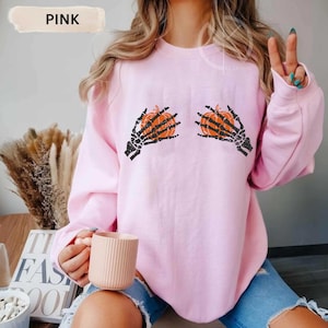 May include: Pink sweatshirt featuring a Halloween-themed design. The graphic depicts two orange pumpkins held by black skeleton hands. The word "PINK" is displayed in a white rectangular box at the top left of the sweatshirt.