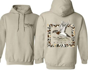 May include: Beige hoodie with a front pocket and drawstring. The front has the word "SITKA". The back features a duck illustration in a camouflage frame, with "SITKA" below. A casual, comfortable garment.