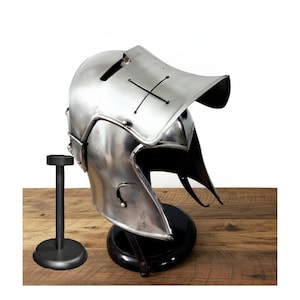 Pode incluir: Um capacete medieval prateado polido com uma viseira distinta e um design de cruz. O capacete é exibido em um suporte preto, mostrando seu design histórico. O capacete é feito de metal e é uma réplica.