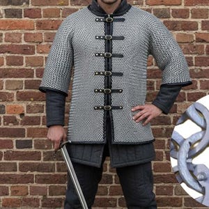 May include: A silver chainmail shirt with black leather accents and closures. The medieval-style armor features short sleeves and is worn over a dark gray padded garment. A sword is held in the hand. The background is a brick wall.