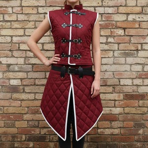 May include: A burgundy quilted vest and skirt set with white trim and black accents. The vest has a high collar and is fastened with black buckles. The skirt is knee-length and features a diamond pattern. A black belt cinches the waist.