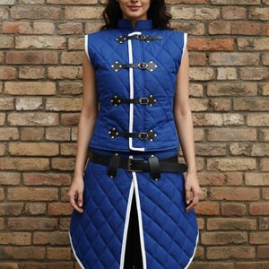 May include: A blue quilted vest and skirt set with white trim and black leather straps. The vest has a high collar and decorative gold rivets. The skirt has a unique, curved hemline and is secured with a black belt.