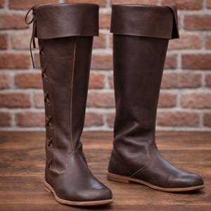 May include: A pair of brown leather knee-high boots. The boots have a folded-over top and are laced up the side. The soles are a lighter brown color. The boots are displayed on a wooden surface in front of a brick wall.